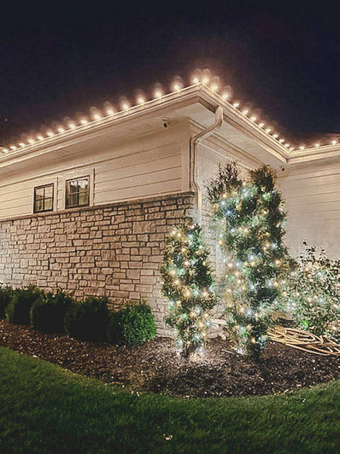 Warm roofline lighting with lit evergreen shrubs along a stone facade